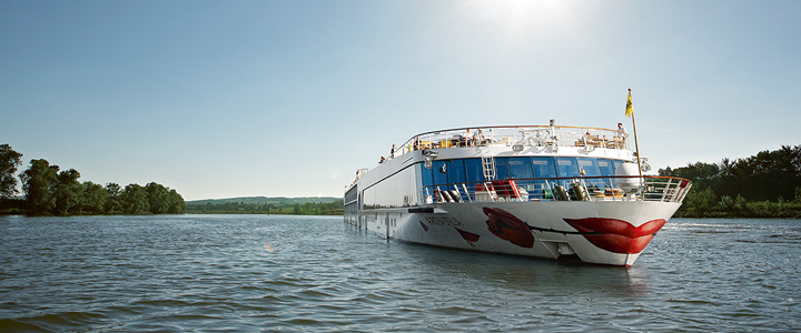 A-Rosa Kreuzfahrtschiff im Fokus, umgeben von ruhigem Gewässer; Bäume im Hintergrund.