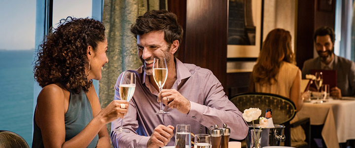 Pärchen an Bord eines Kreuzfahrtschiffes im Restaurant. Beide stoßen lächelnd mit einem Glas Sekt an.